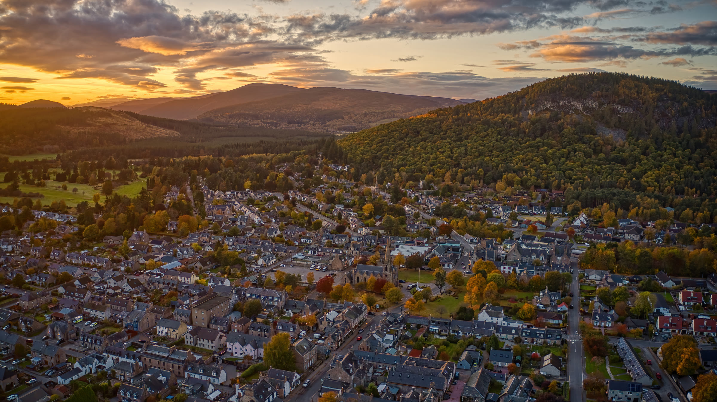 Ballater from the air at an autumn sunset, its grey granite streets and the church on its green below the wooded hill of Craigendarroch, the hills of Deeside beyond.