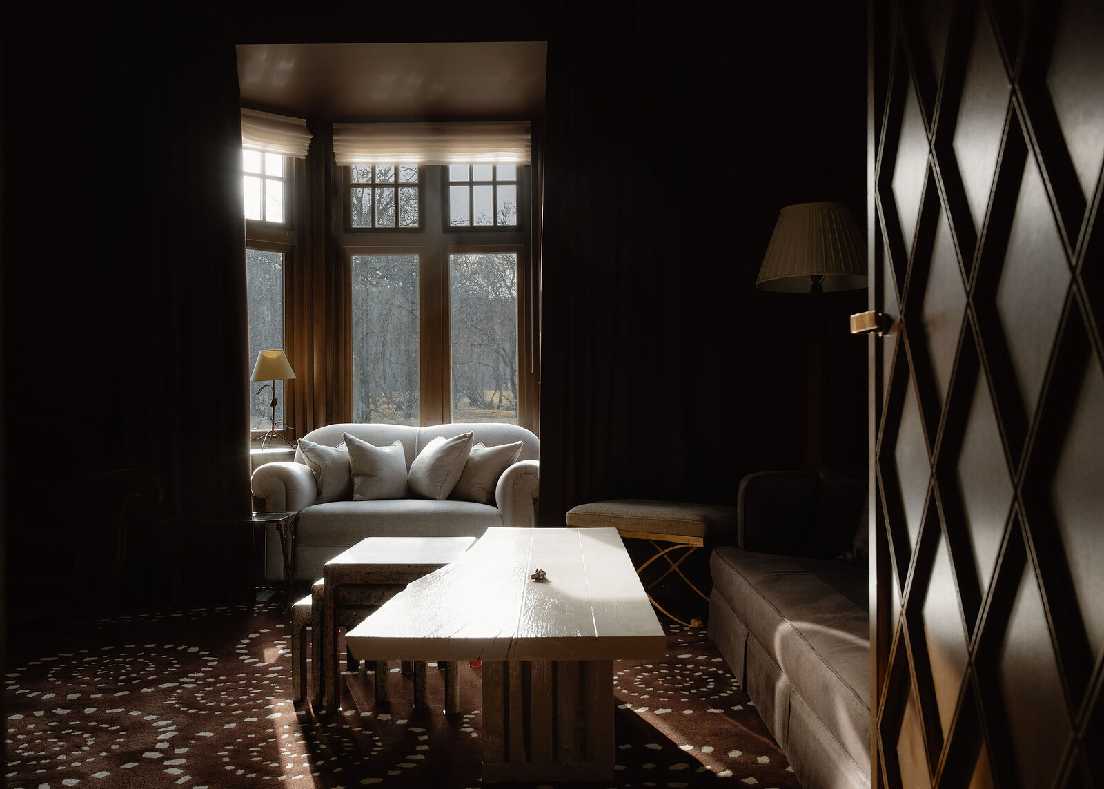 A landscape view of the dark-panelled lounge: sofas round a white timber table under the bay window, a door carved with diamonds at the side.