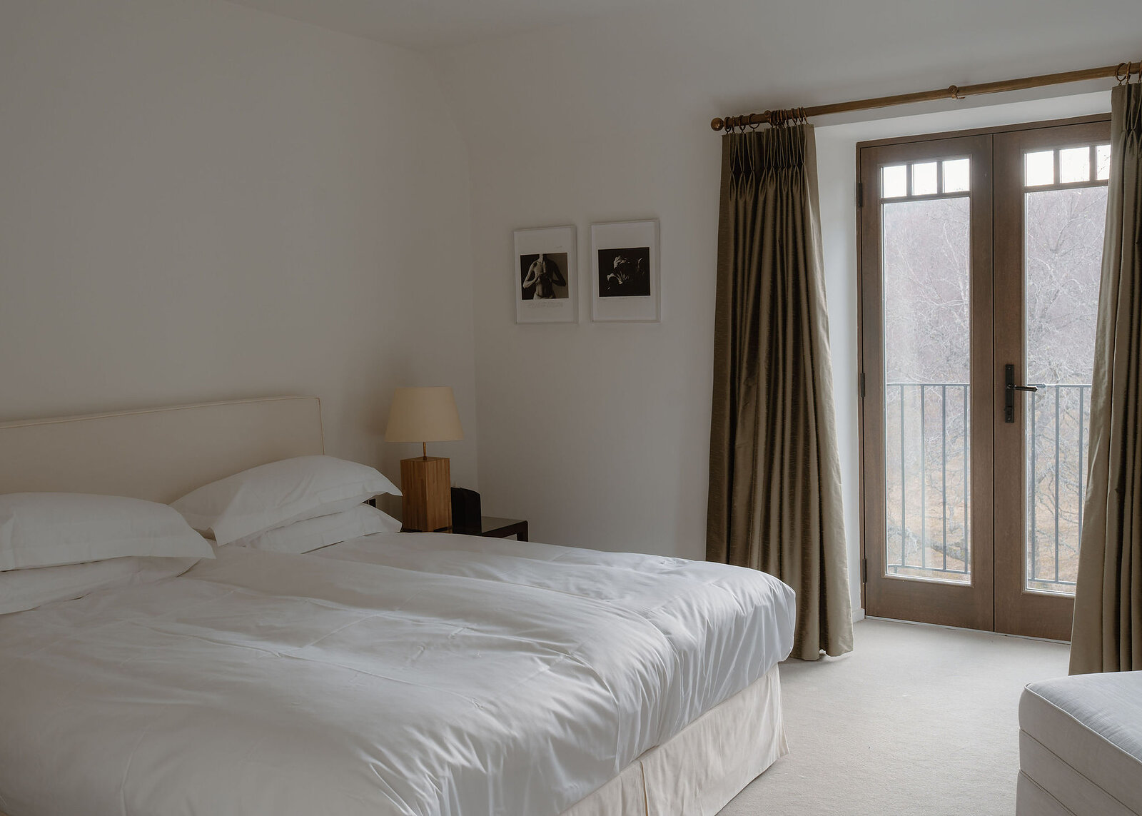 A landscape view of the bedroom: the bed made up in white, olive curtains at the French doors and a footstool.