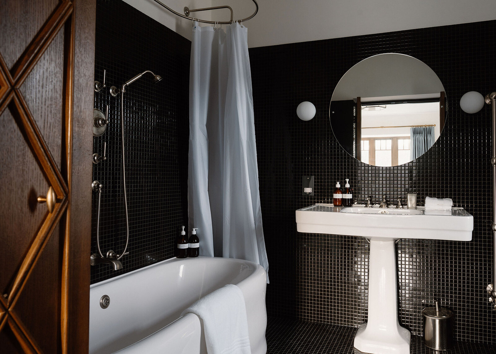 A landscape frame of the bath, its curtain rail and shower, the pedestal basin and globe lights on the black mosaic.