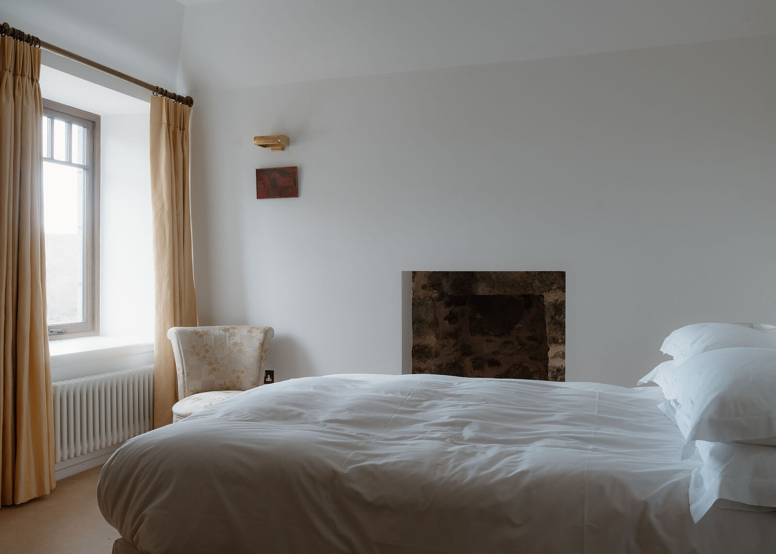 A landscape view of a bedroom: the white bed, the stone fireplace opening, a floral armchair and golden curtains at the window.