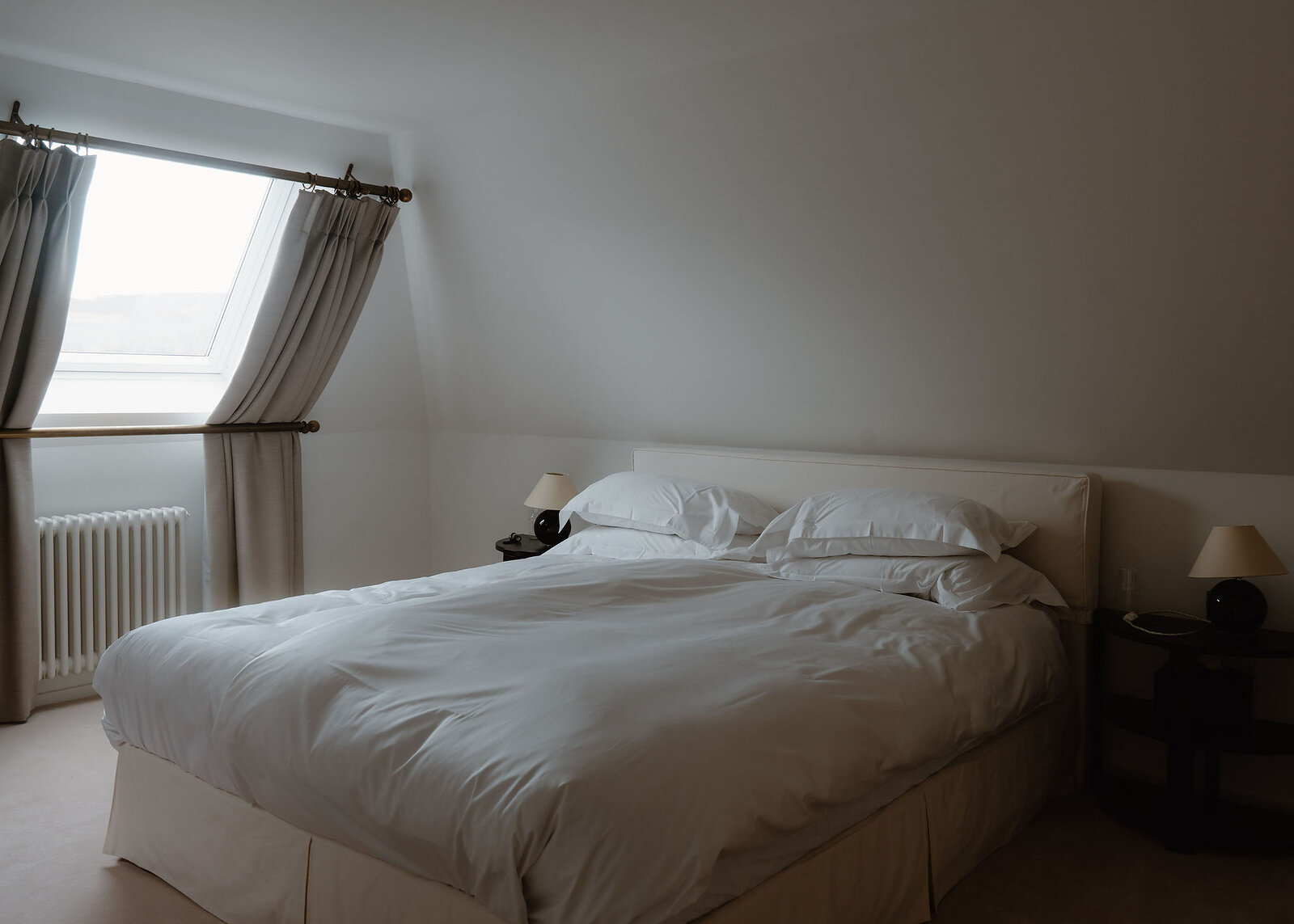 A landscape view of the attic bedroom: the bed, black lamps on the side tables and a roof window with curtains.