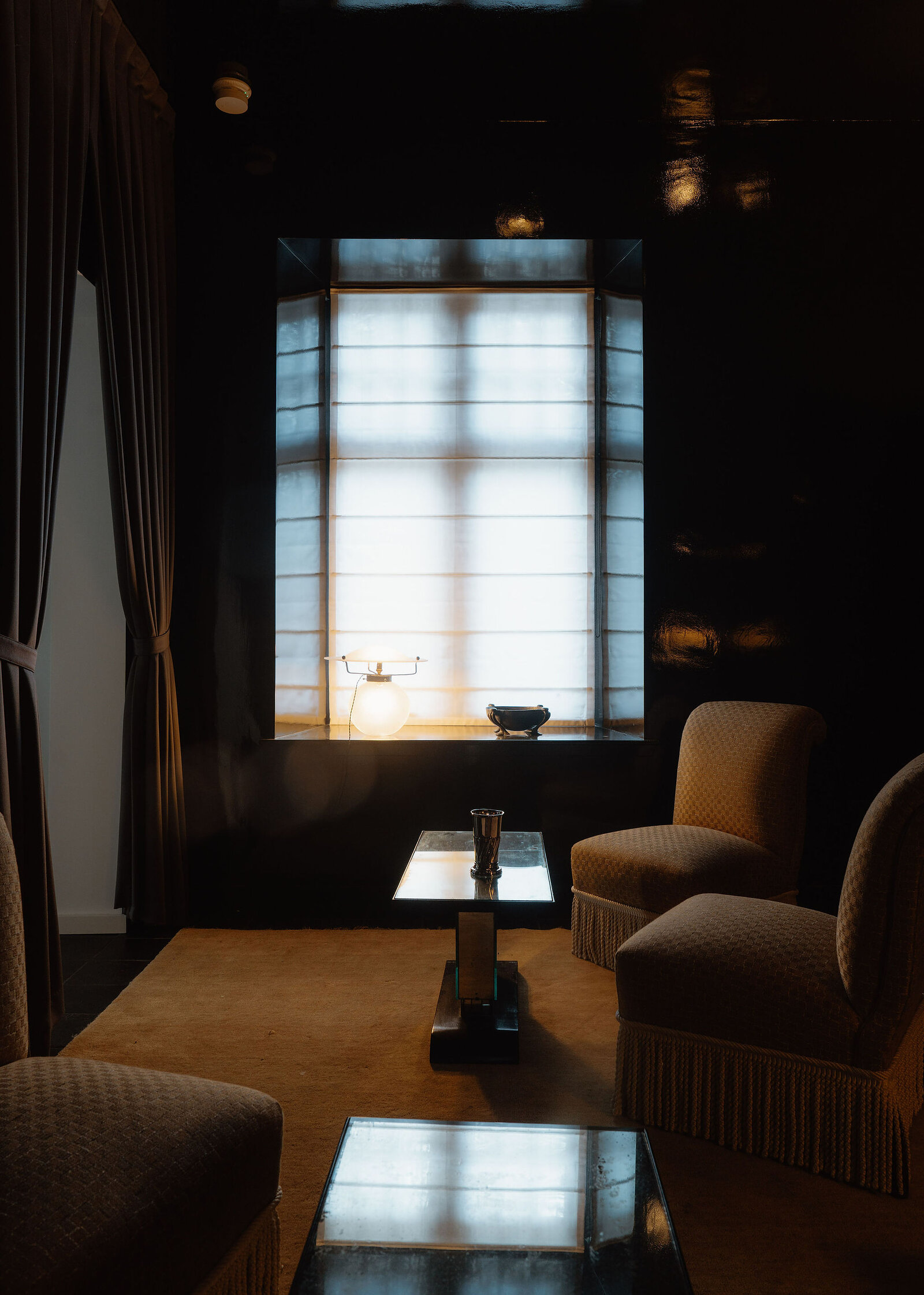 A dark sitting room with fringed armchairs round a glass table, light coming through a leaded window.