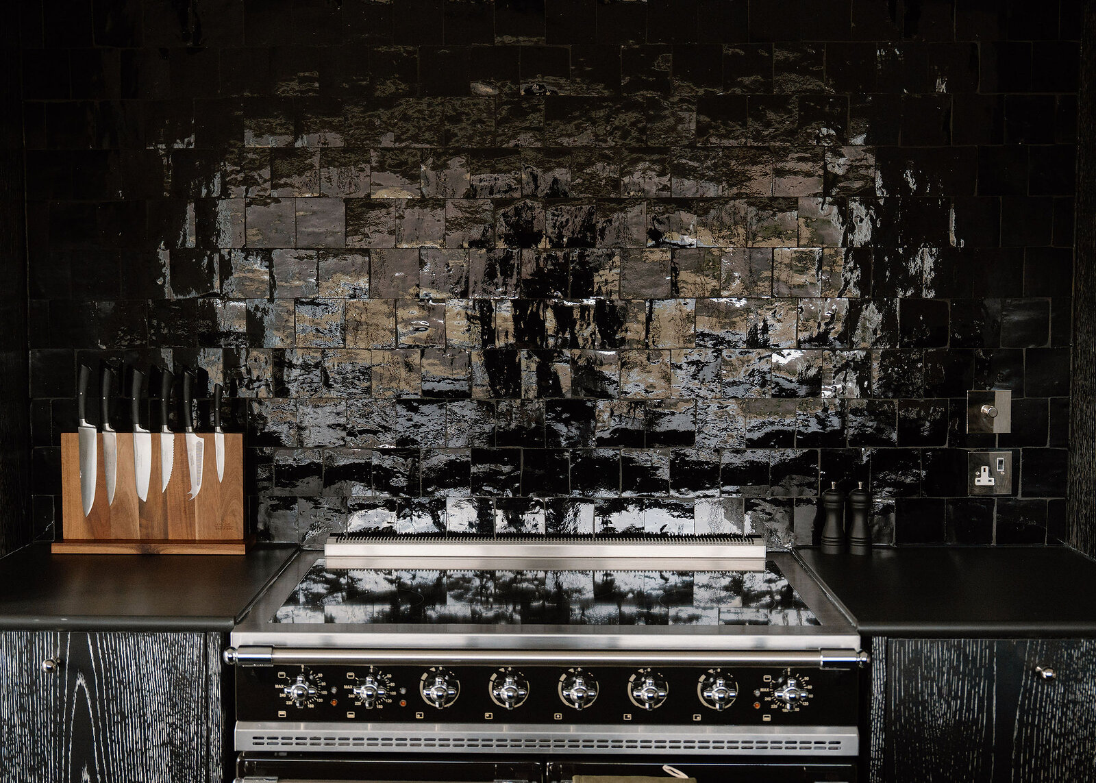 A black range cooker against a wall of glossy black handmade tiles, a block of knives on the worktop.