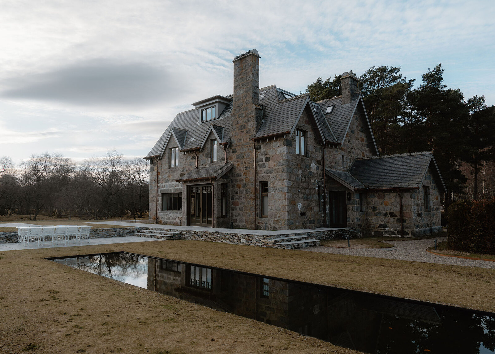 A landscape view of Faunoran House from the garden, the terrace and its white table, the house reflected in the long pond.