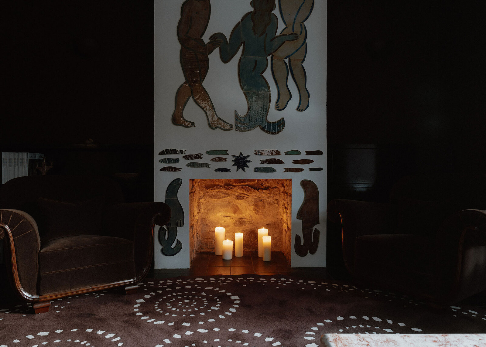 A landscape view of the artist-painted fireplace between two curved velvet armchairs, candles in the hearth.