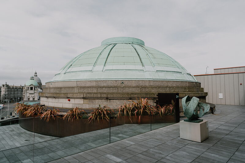 A glazed dome rising above a rooftop terrace, a bronze sculpture standing beside planters along the railing.