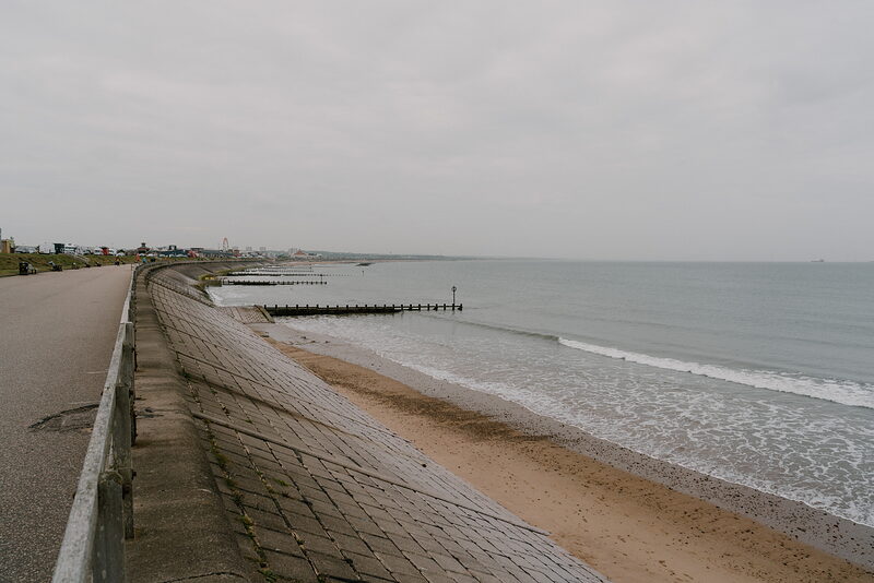 A promenade running beside a rocky breakwater and sandy beach, waves breaking along the shore.