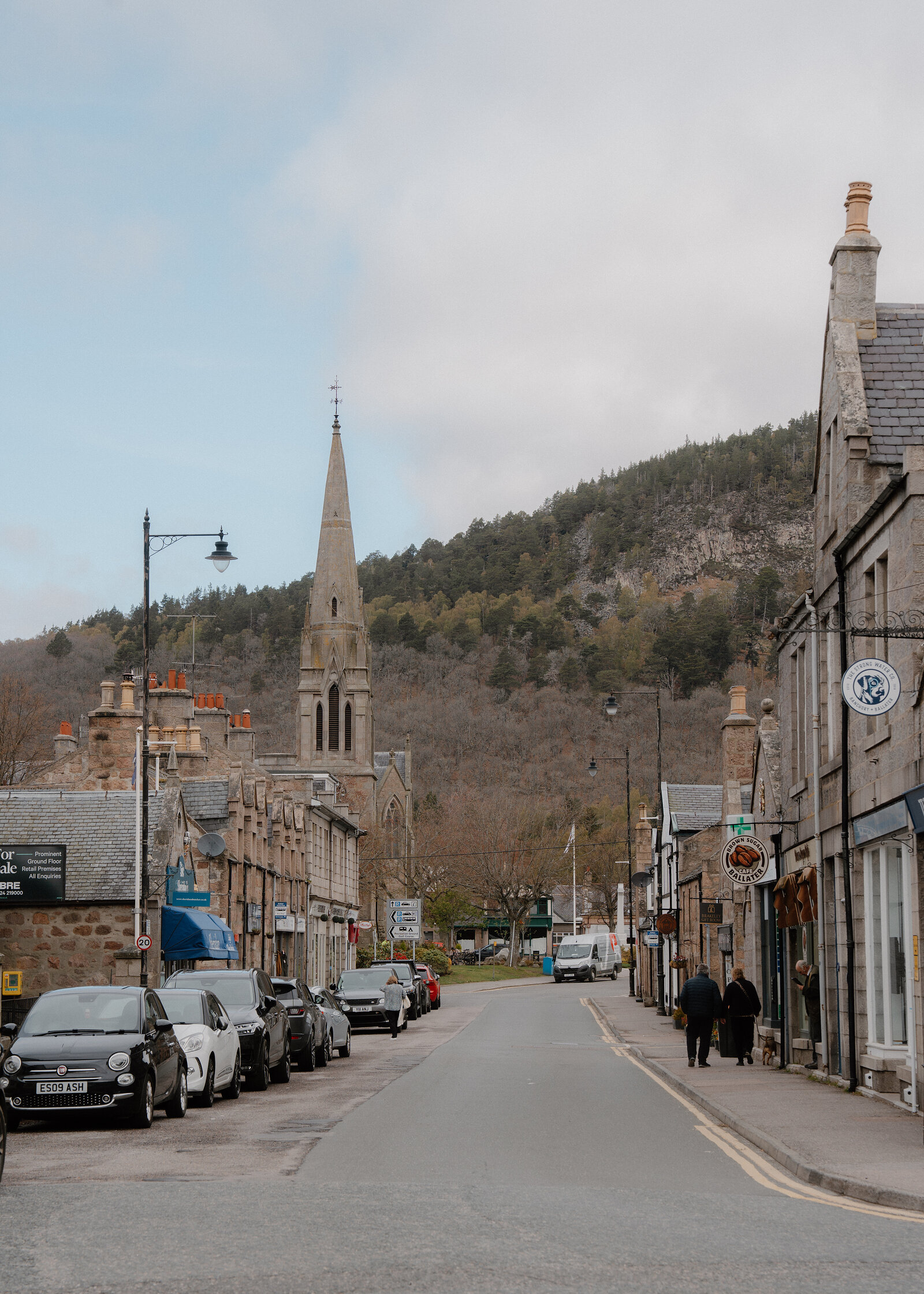 A church spire rises above Ballater's high street, shoppers walking past parked cars and hillside beyond