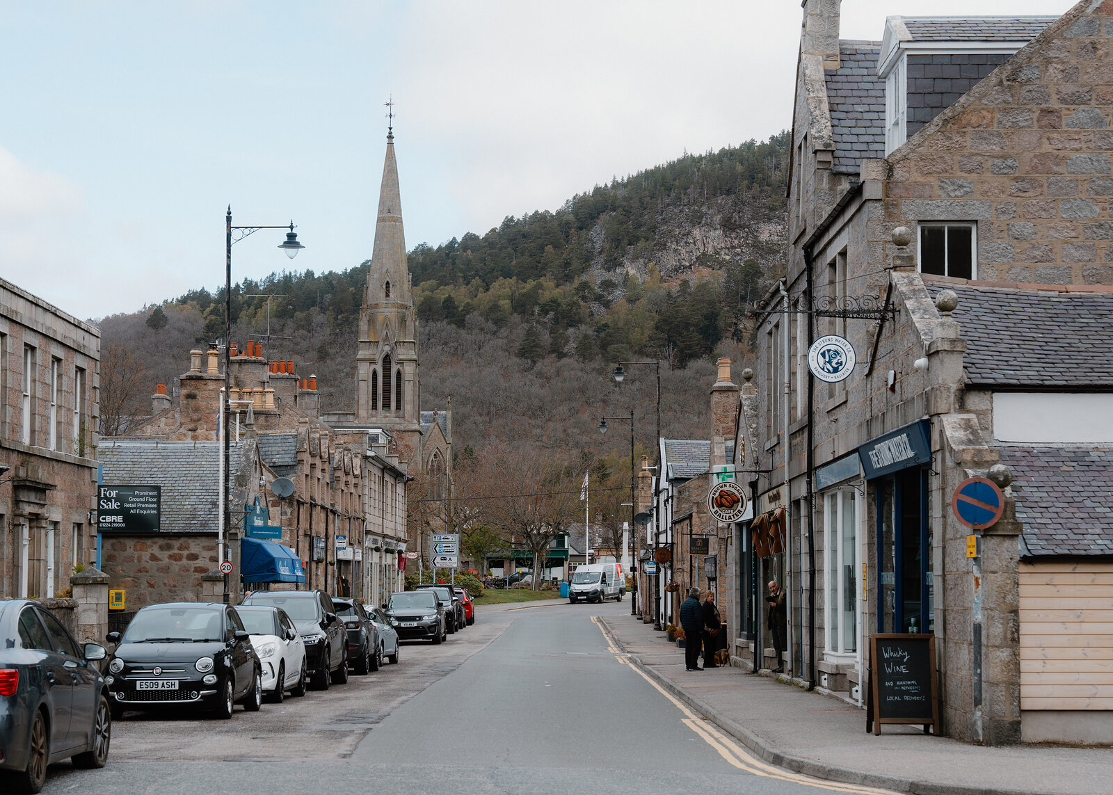 Ballater's high street runs towards a church spire beneath wooded hills, shoppers passing the storefronts