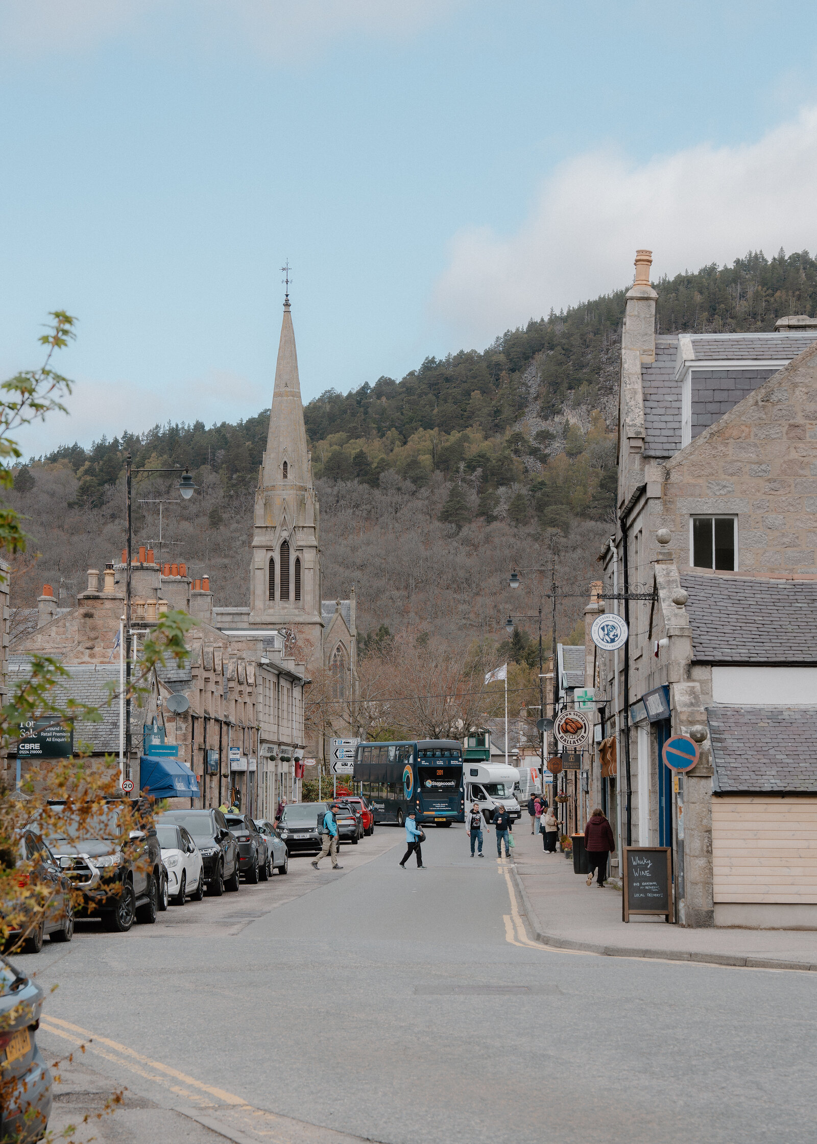 Pedestrians cross Ballater's high street as a bus passes beneath a church spire and wooded hills