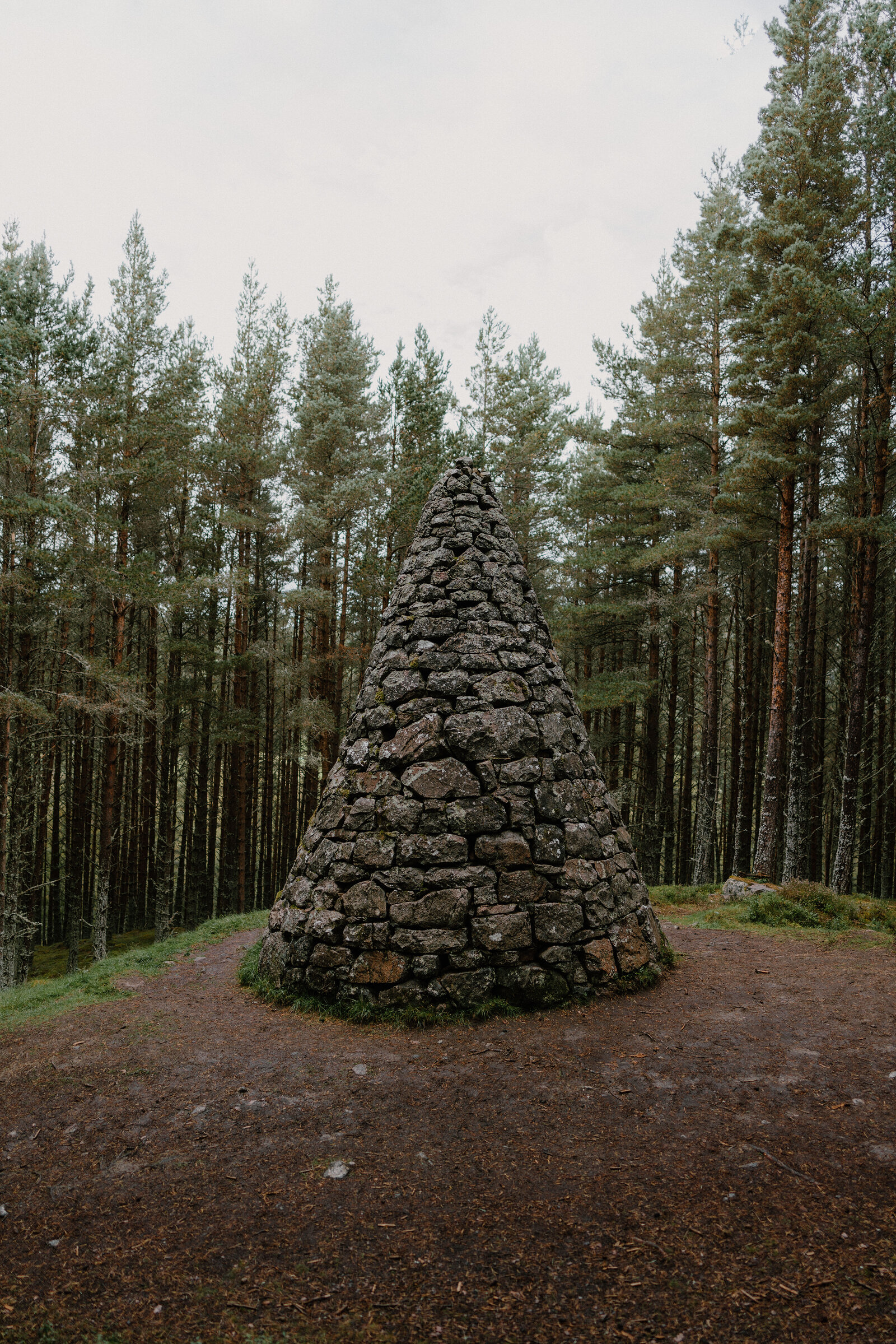 A stone cairn built into a cone shape, standing in a clearing among pine trees