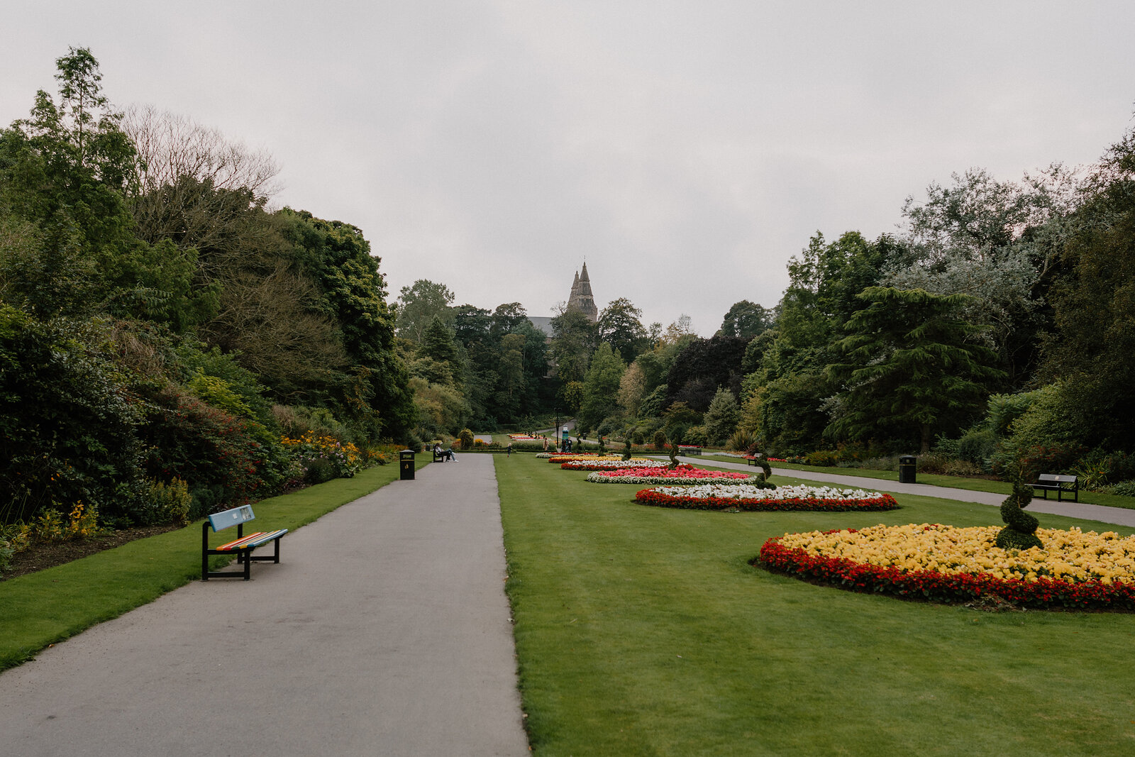 A formal park path lined with flower beds and clipped hedges, a spire visible in the distance