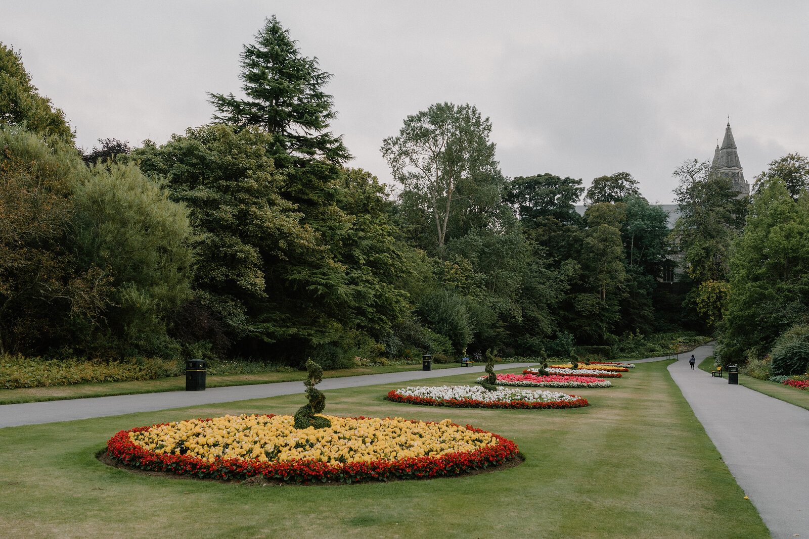 A formal park path lined with flower beds and clipped hedges, a figure walking in the distance