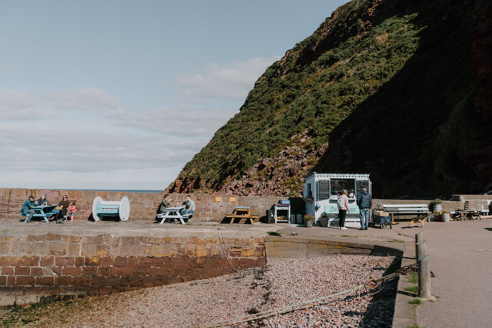 People seated at picnic tables beside a food van below a green cliff