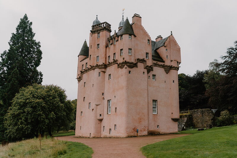 A pink turreted castle standing behind trees, a stone wall bordering the lawn.