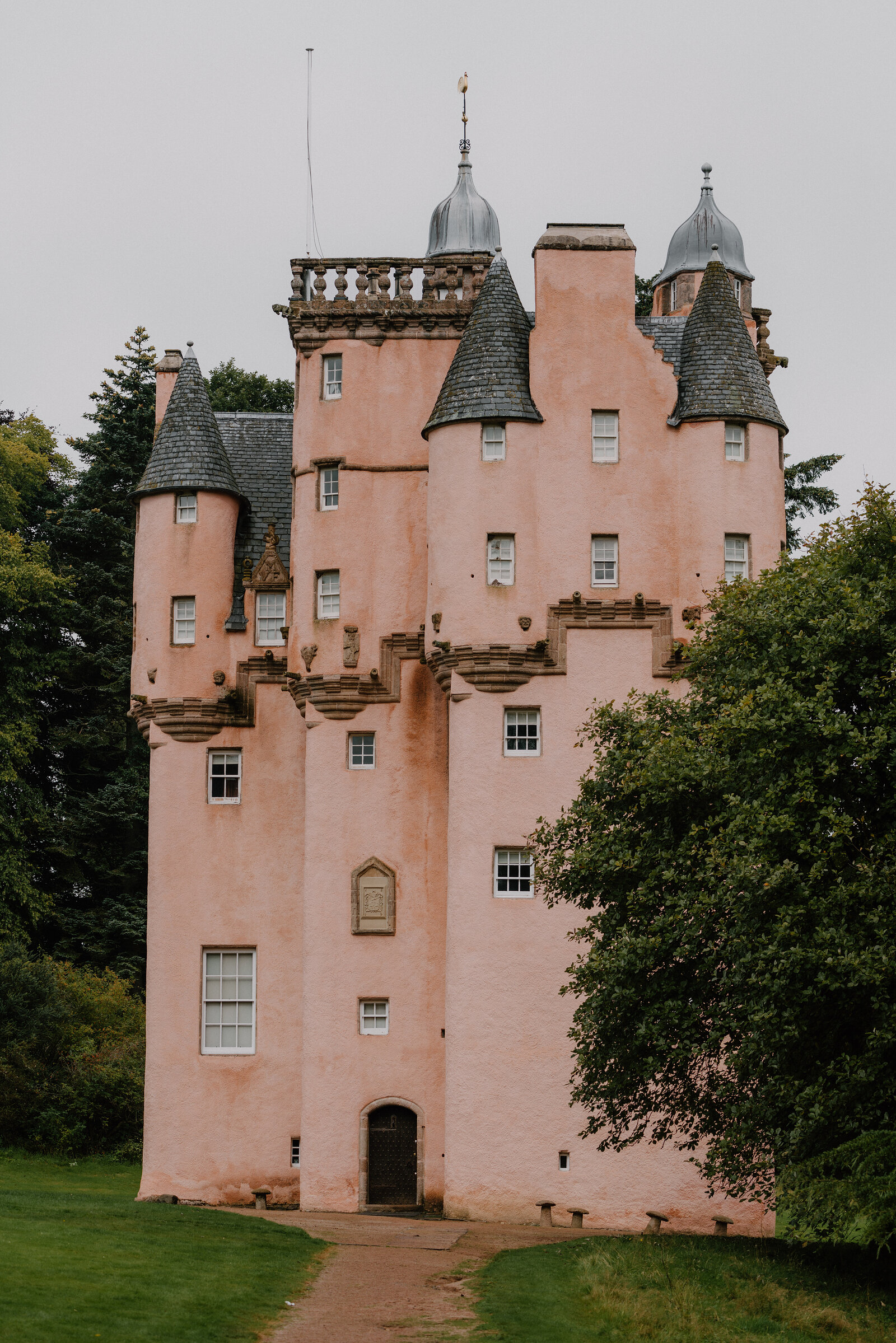 A pink turreted castle standing among trees, its stepped gables catching the grey light