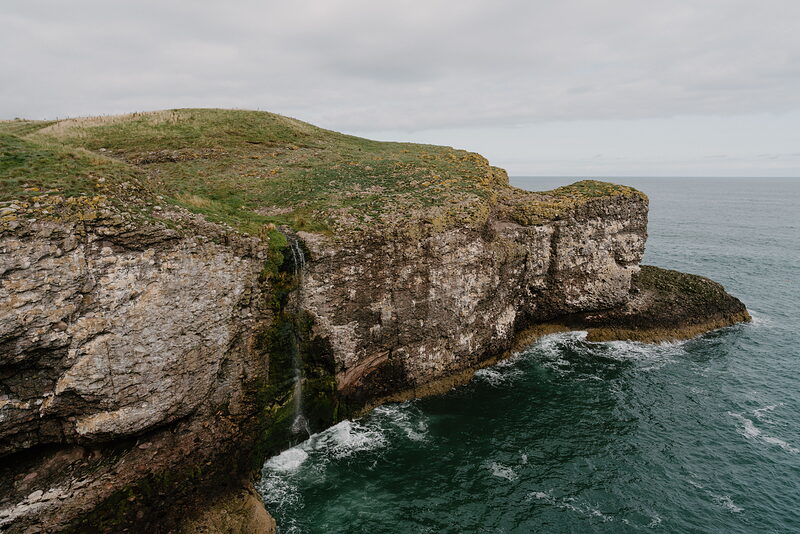A grassy headland with a small waterfall spilling into the sea, cliffs rising on either side.
