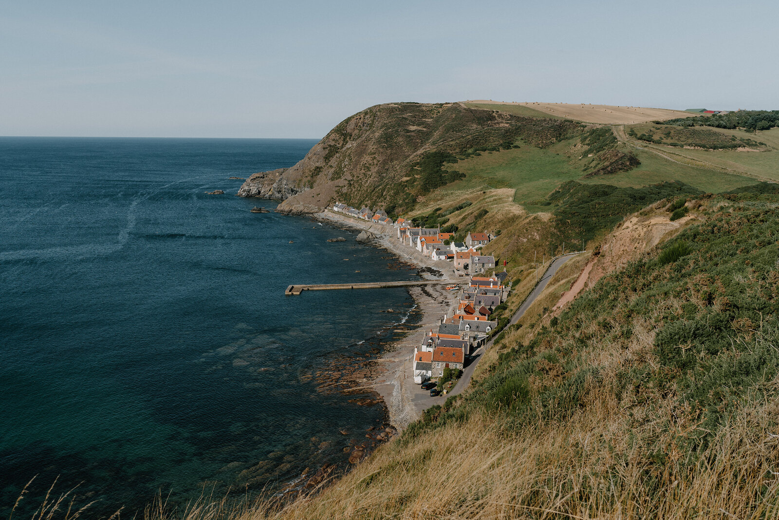 A fishing village strung along the shoreline beneath a grassy headland, a harbour wall reaching into the sea
