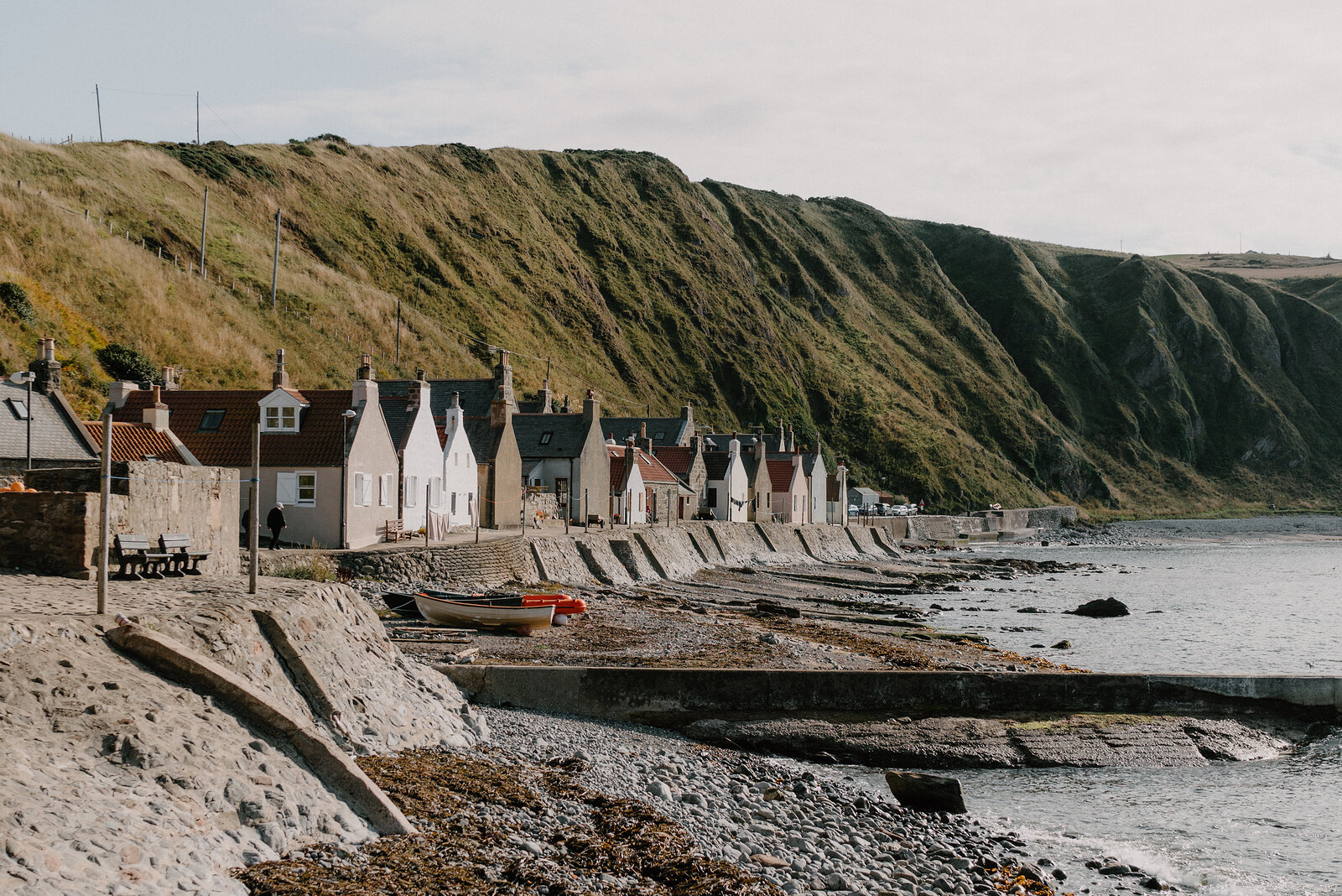A row of cottages strung along the base of a grassy cliff, a small boat resting on the shore