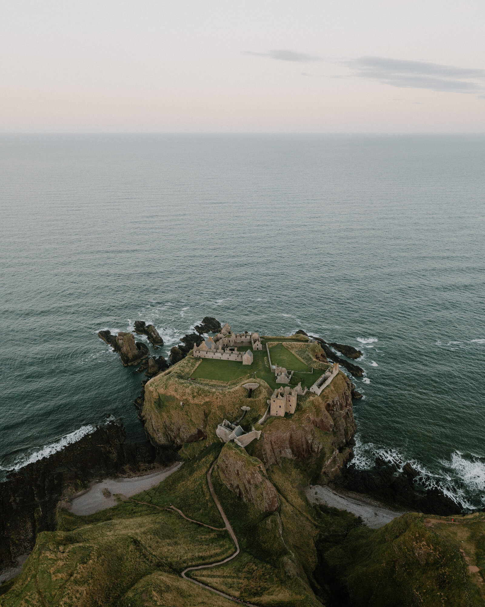 A ruined castle standing on a rocky headland surrounded by the sea, a path winding up from the cliffs