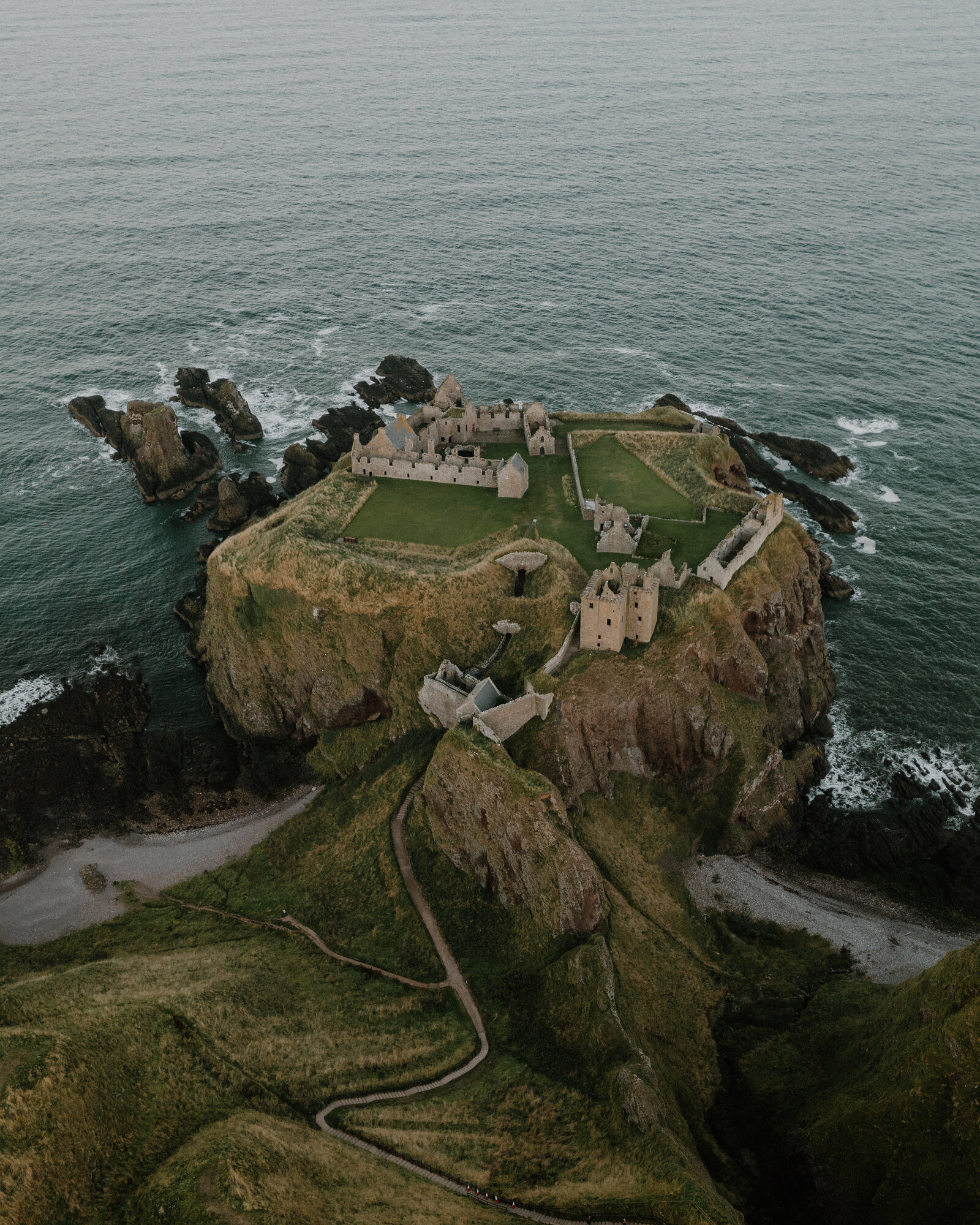 A ruined castle standing on a grassy headland surrounded by the sea, seen from above