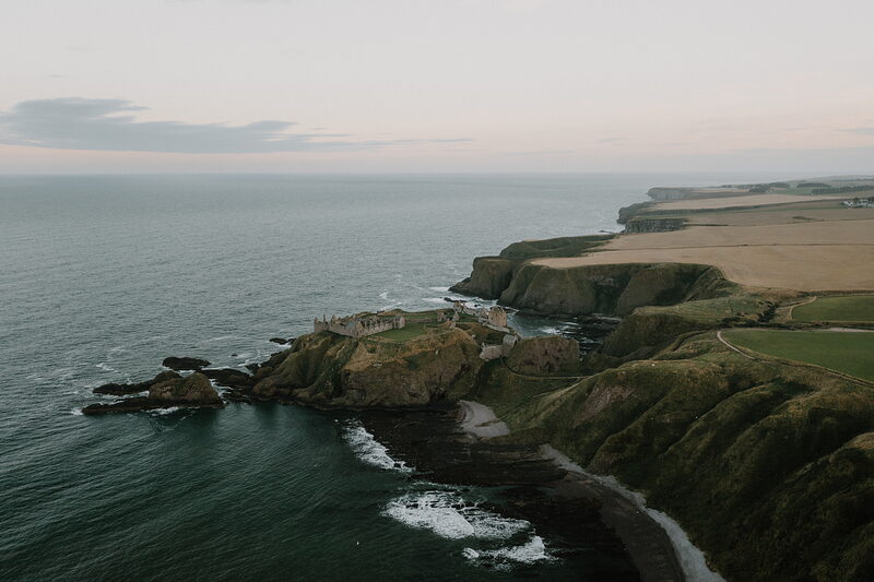 A ruined castle standing on a rocky headland surrounded by the sea, fields stretching along the clifftop.