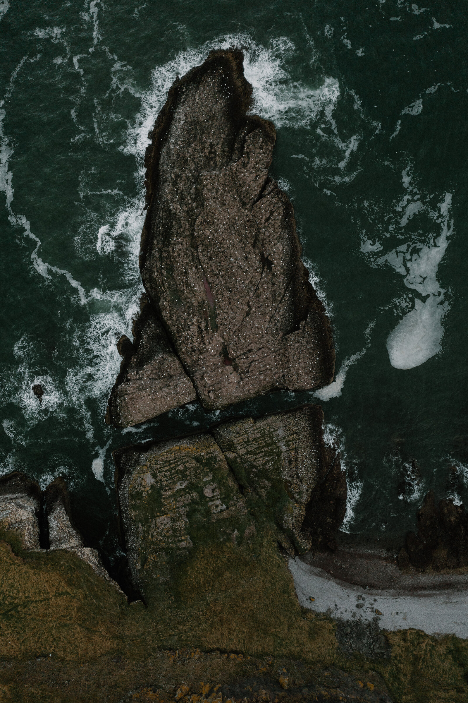A rocky sea stack surrounded by breaking waves, seen from directly above