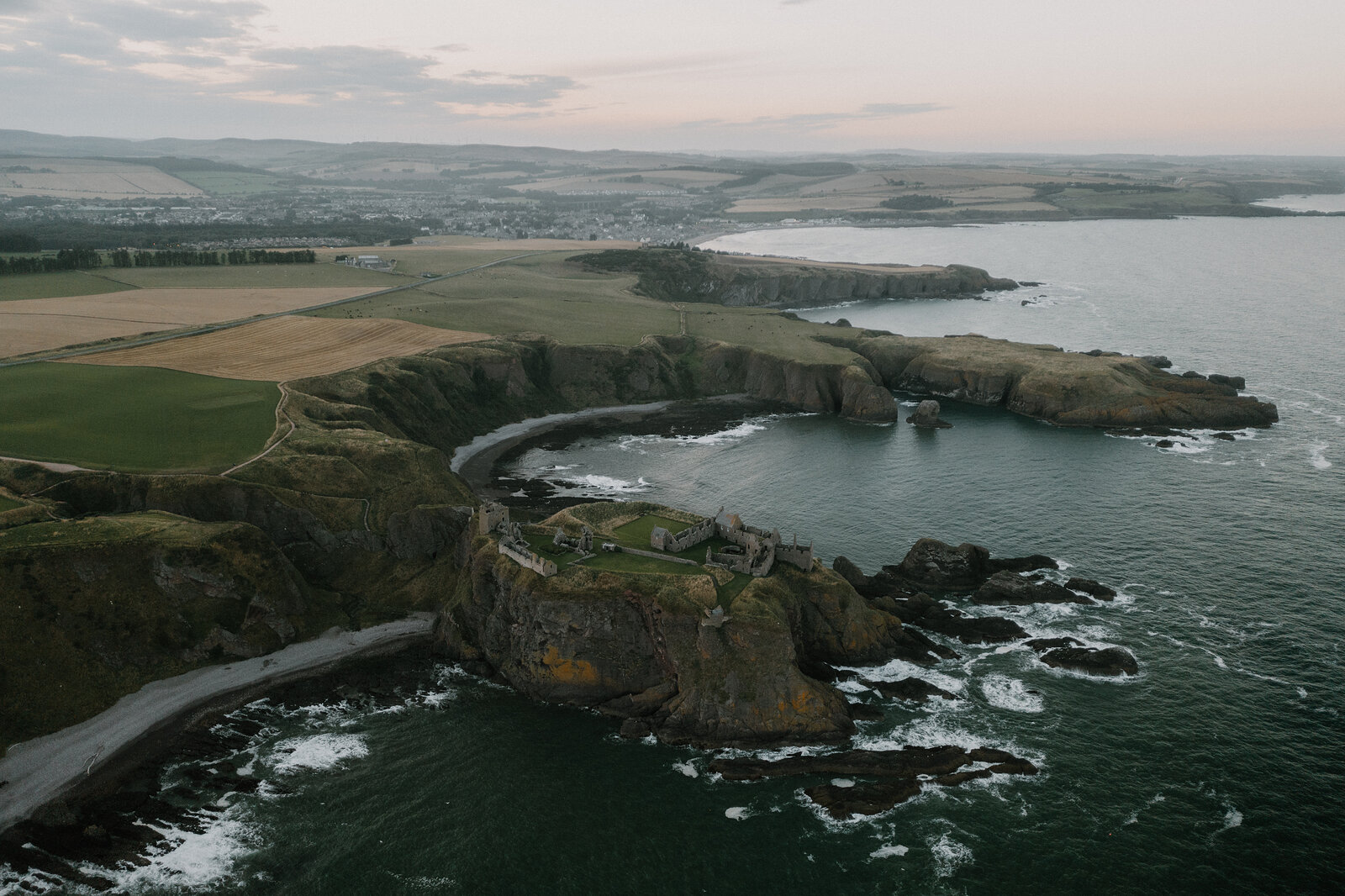 An aerial view of a ruined castle on a rocky headland, farmland and a town stretching along the coast