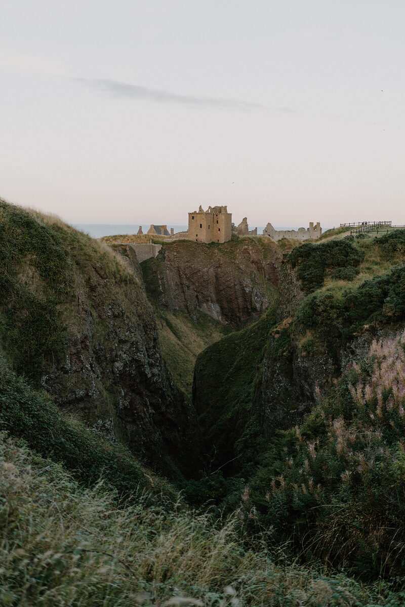 Ruined castle walls crown a sea-cliff promontory beyond a deep grassy chasm.