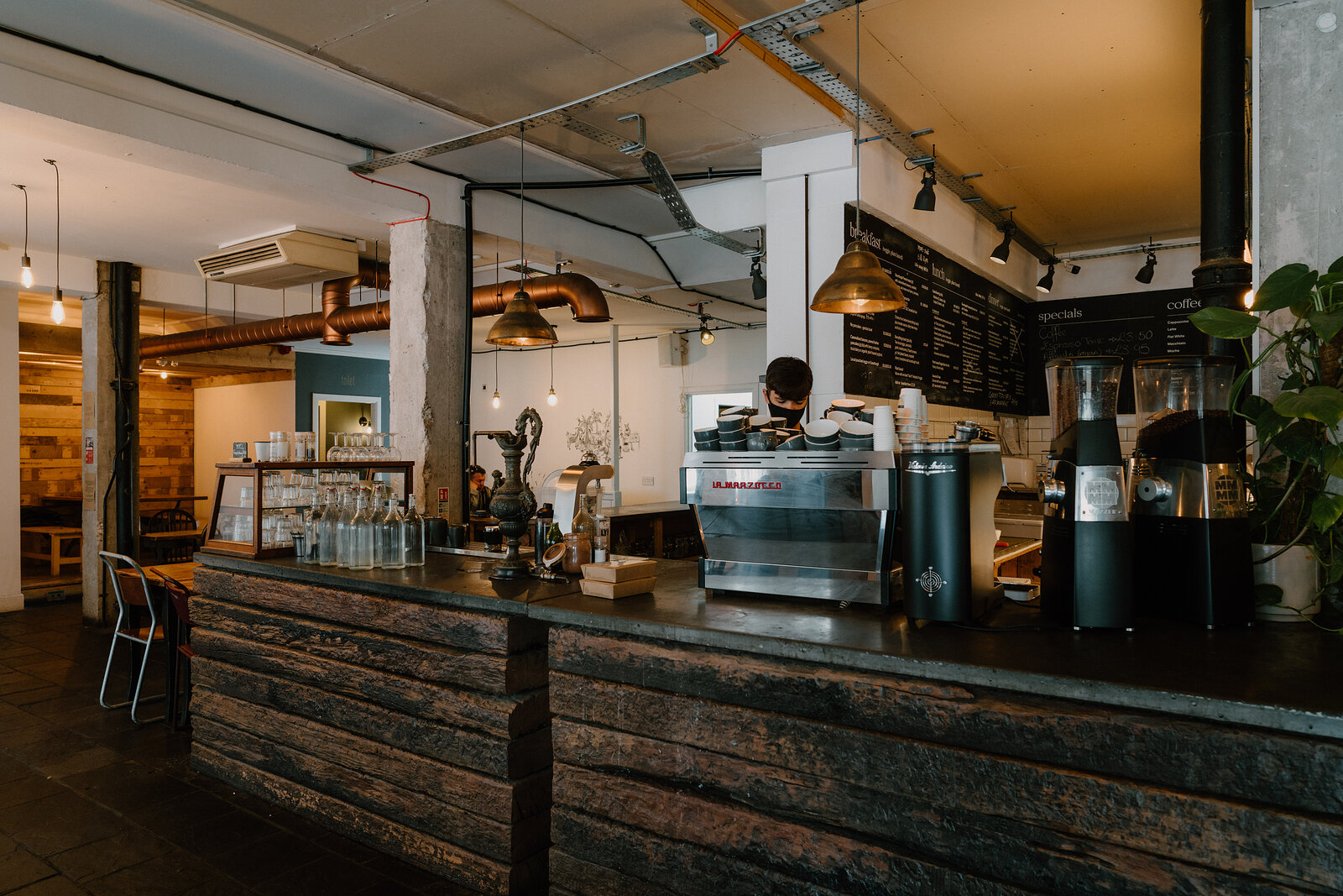 A barista tends an espresso machine beneath hanging bulbs and copper pipework at a bar counter.