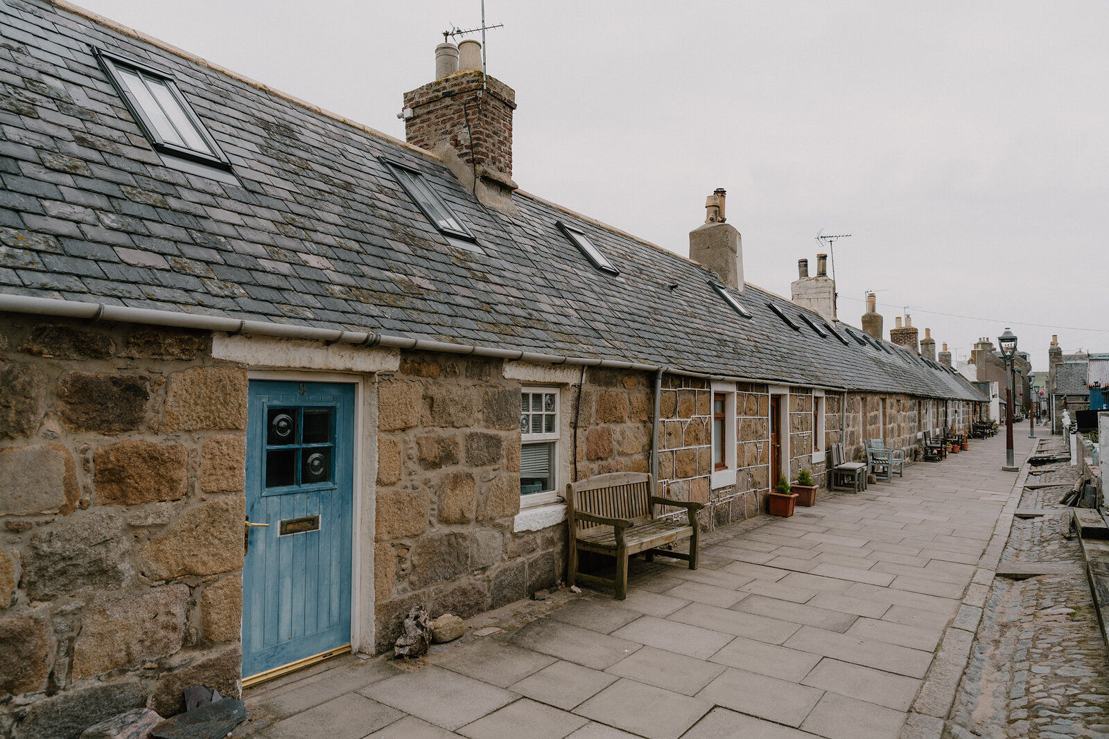 A row of stone cottages with dormer windows lines a cobbled street, a blue door among them.