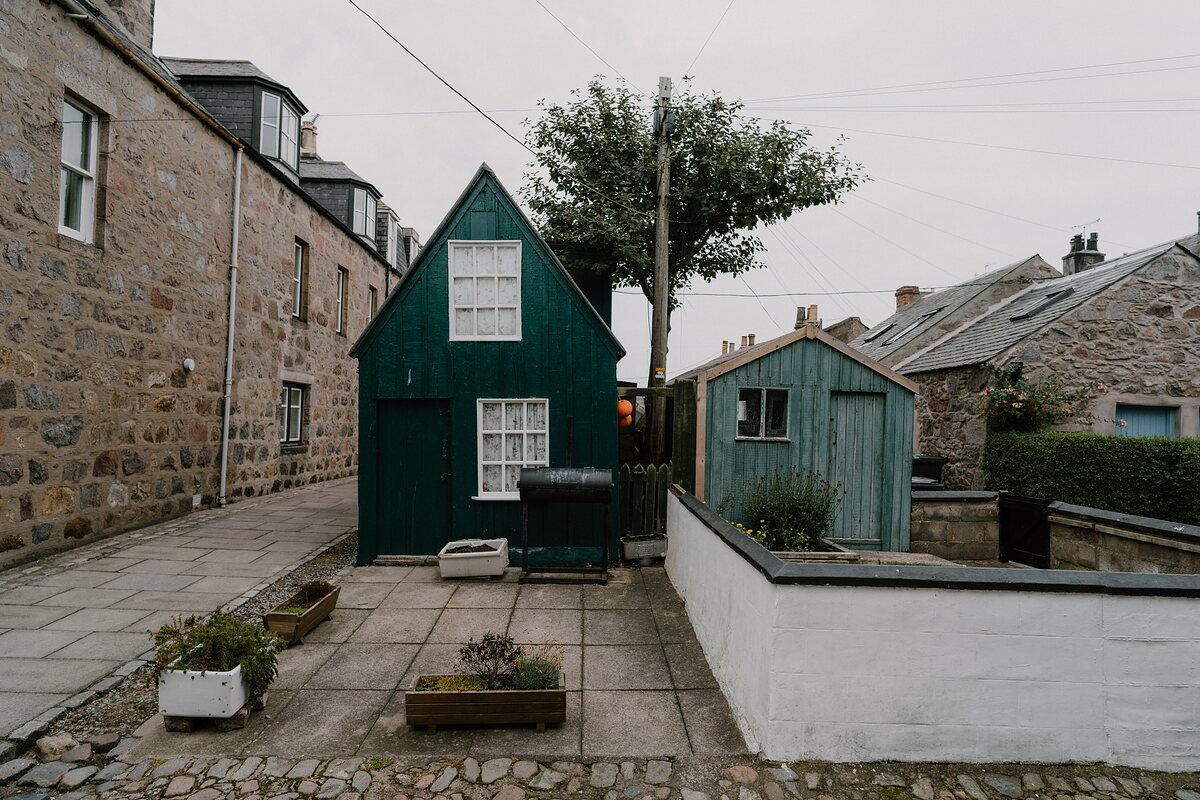 A dark green cottage and a weathered blue shed sit either side of a paved courtyard.