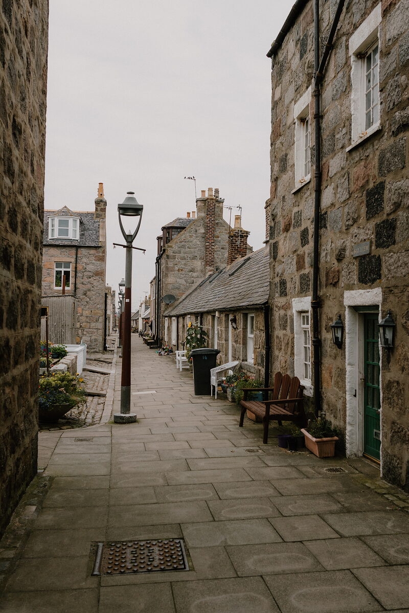 A cobbled street lined with stone cottages leads past a lamppost, bench and potted flowers.