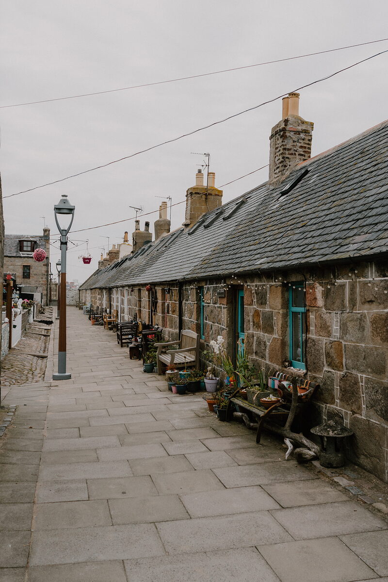 Benches and potted flowers line a cobbled street of stone cottages with teal doors.