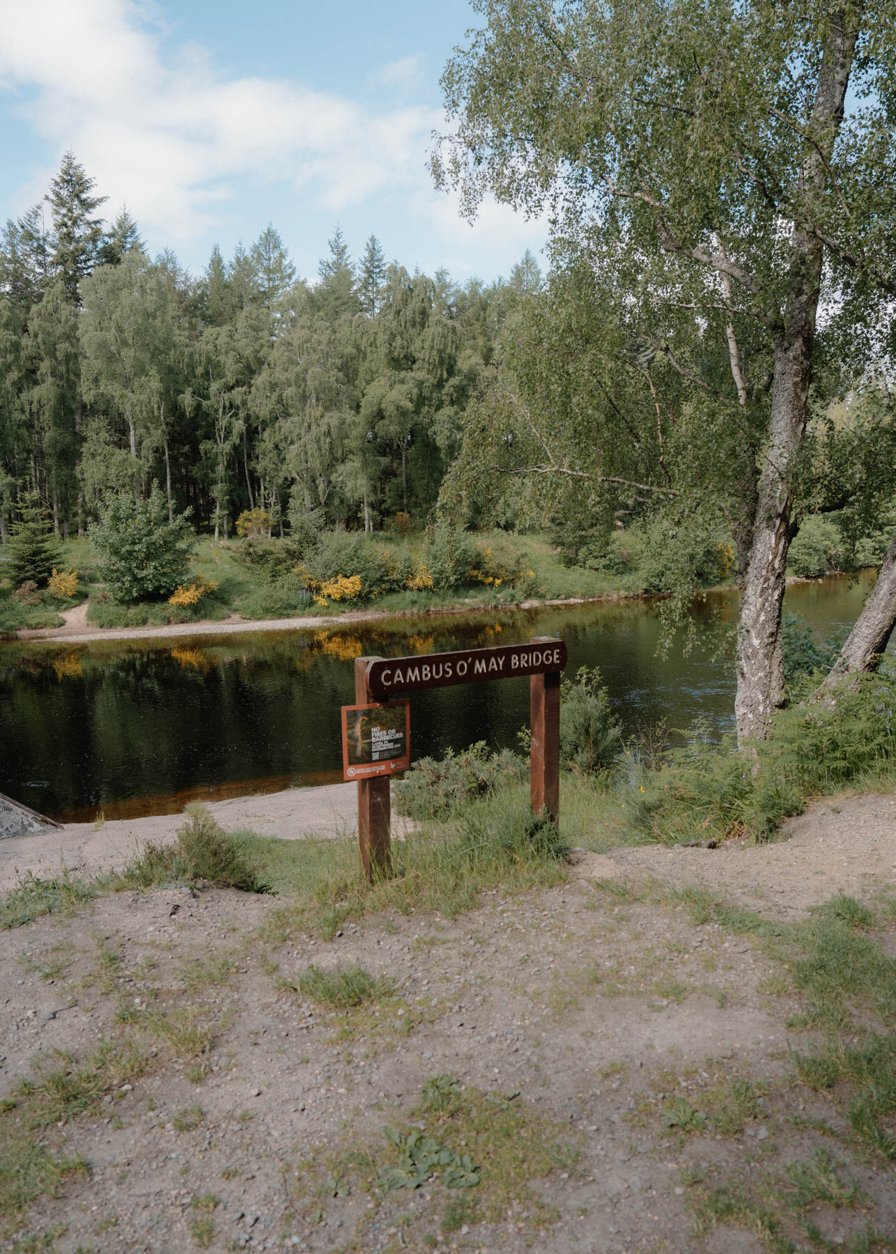 A wooden sign reading Cambus O'May Bridge set beside a calm river bordered by trees