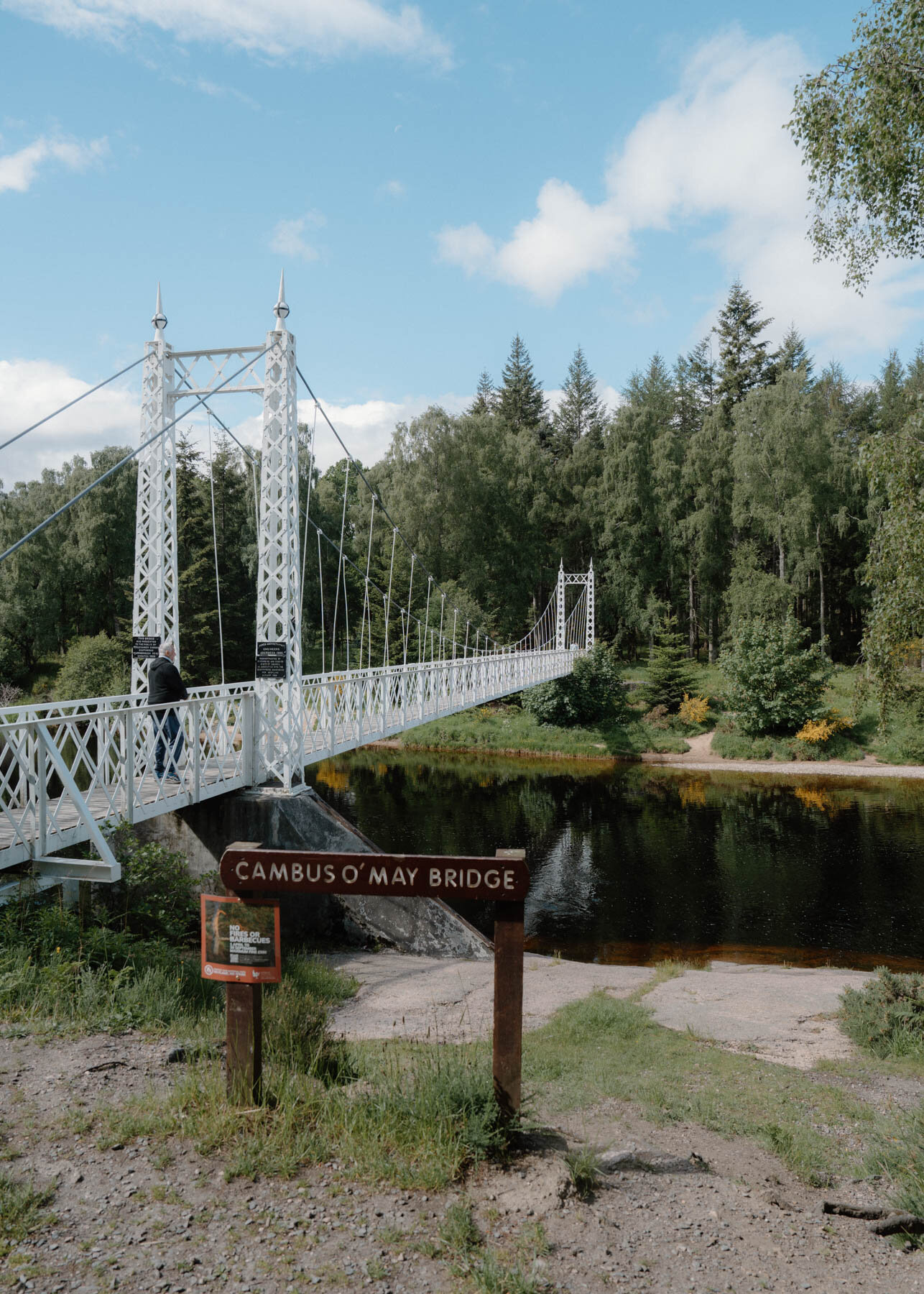 A white suspension footbridge crossing a calm river, a sign reading Cambus O'May Bridge mounted at its foot