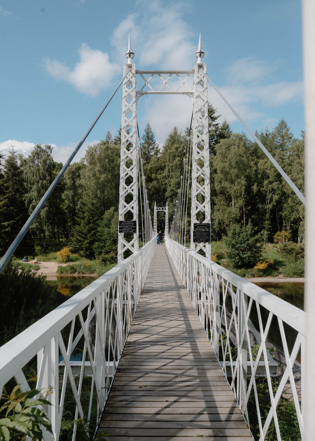 A white suspension footbridge crossing a river, a distant figure crossing near its centre