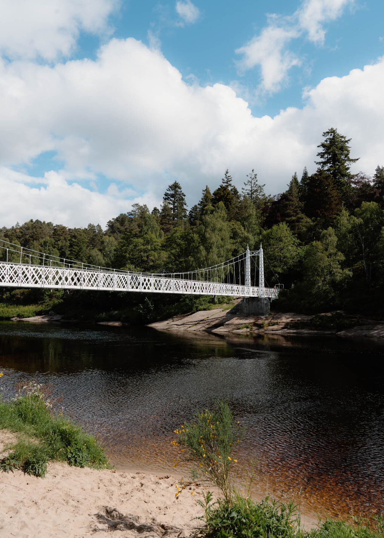 A white suspension footbridge crossing a river bordered by conifers, a sandy bank in the foreground
