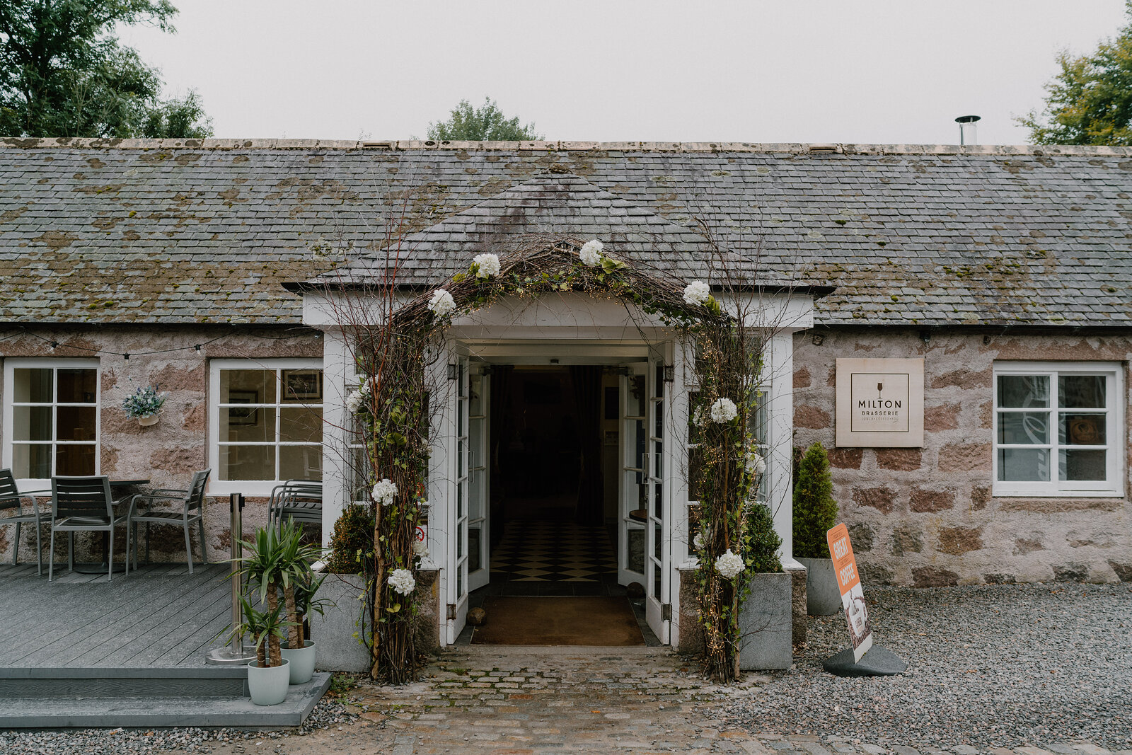 A floral archway frames the entrance to Milton's stone building beside outdoor tables.