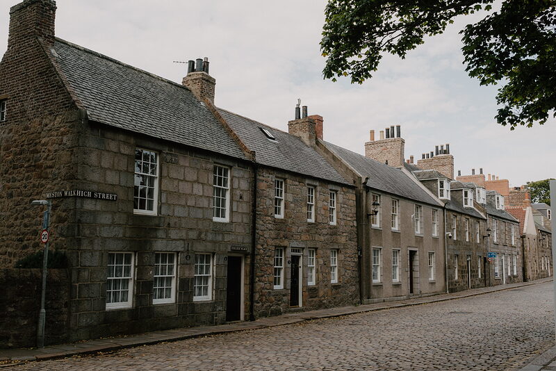 A cobbled Aberdeenshire street lined with granite tenements, a street name plaque mounted on the corner.