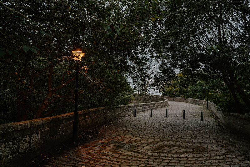 A cobbled path over a bridge in Aberdeenshire, a lit lamppost glowing beneath overhanging trees at dusk.