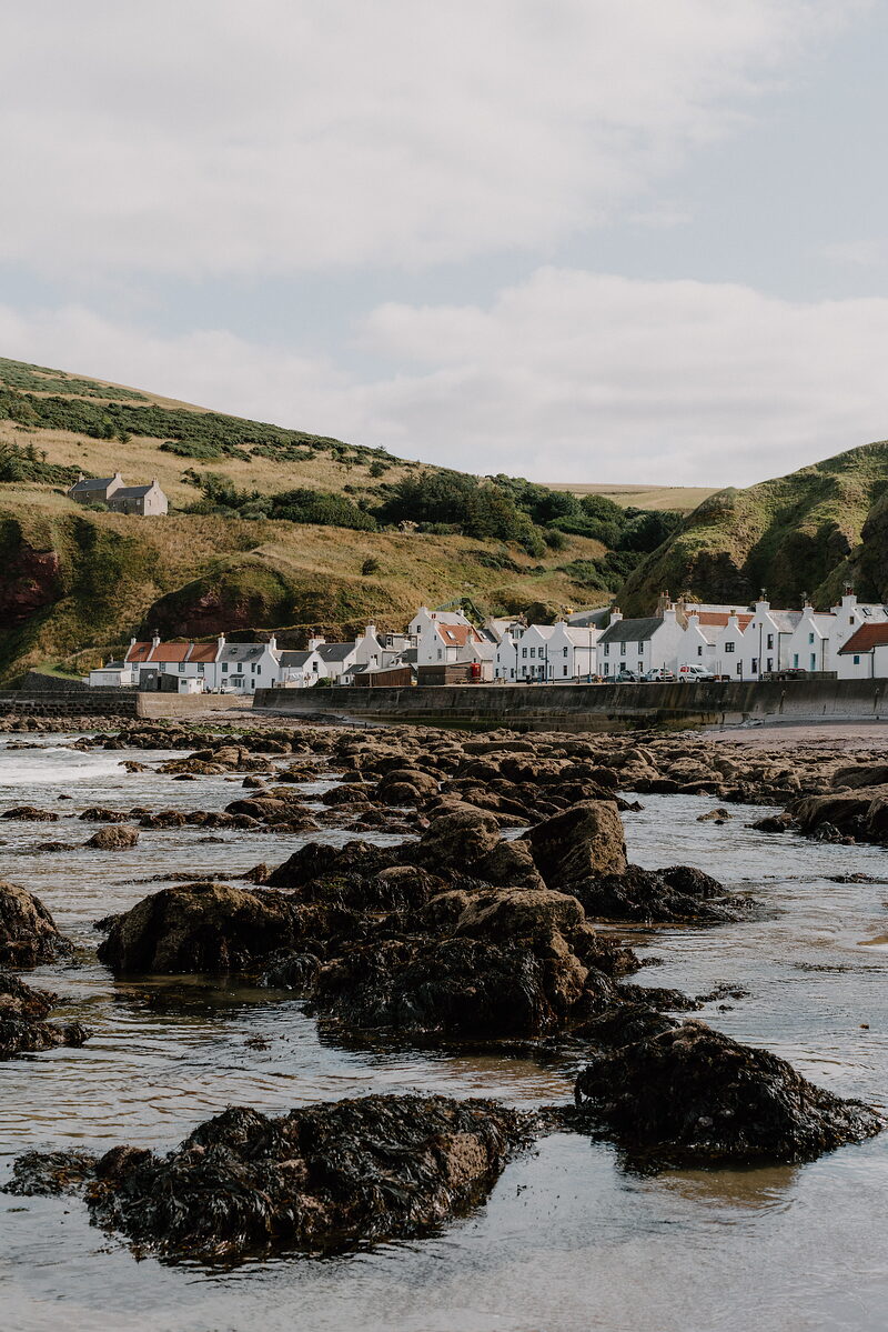 White-harled cottages line the shore of a small Aberdeenshire village beneath green hillsides, seaweed-covered rocks exposed at low tide.