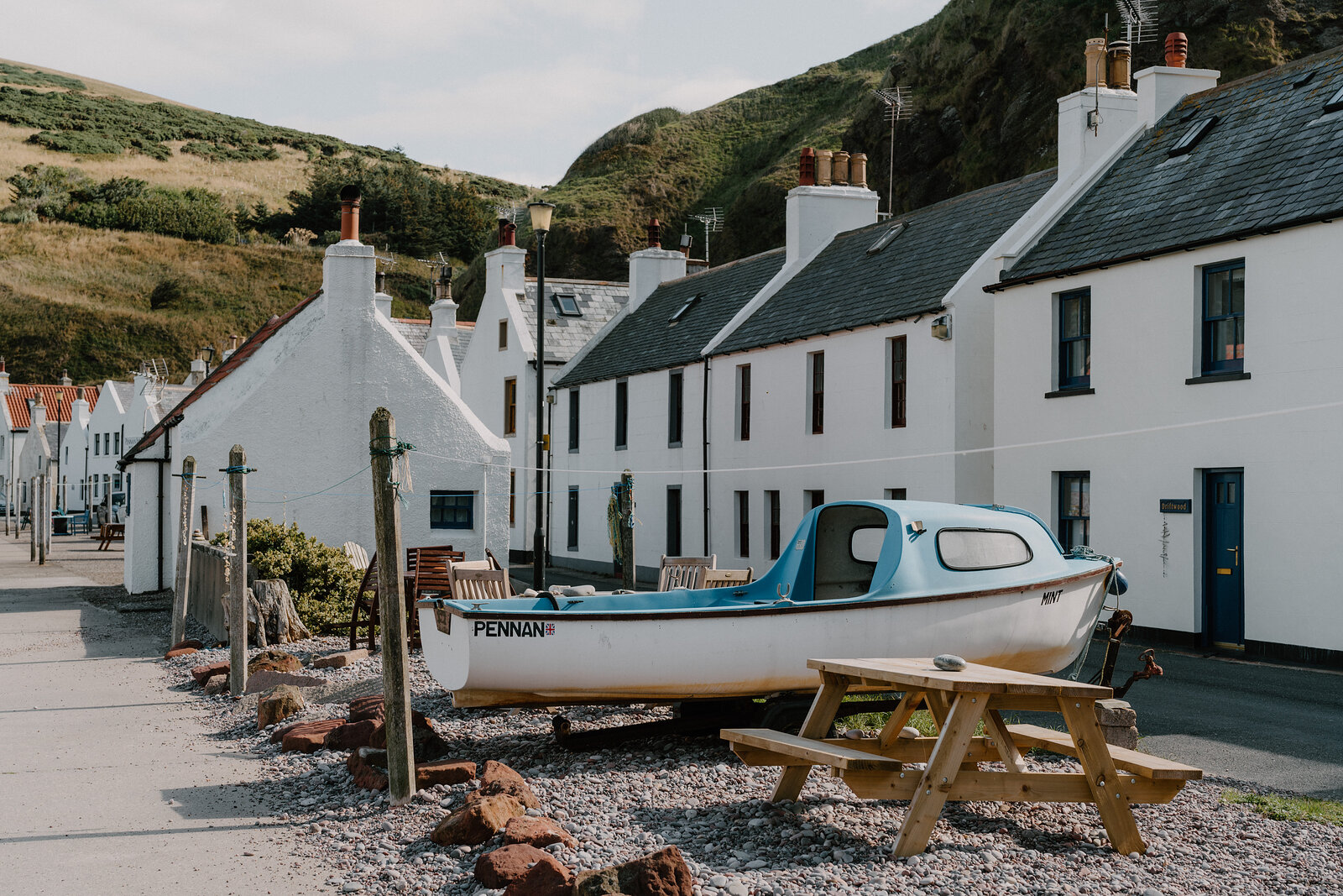 A boat named 'Pennan' rests on the shingle beach in front of the village's white-harled cottages, a picnic table set nearby.