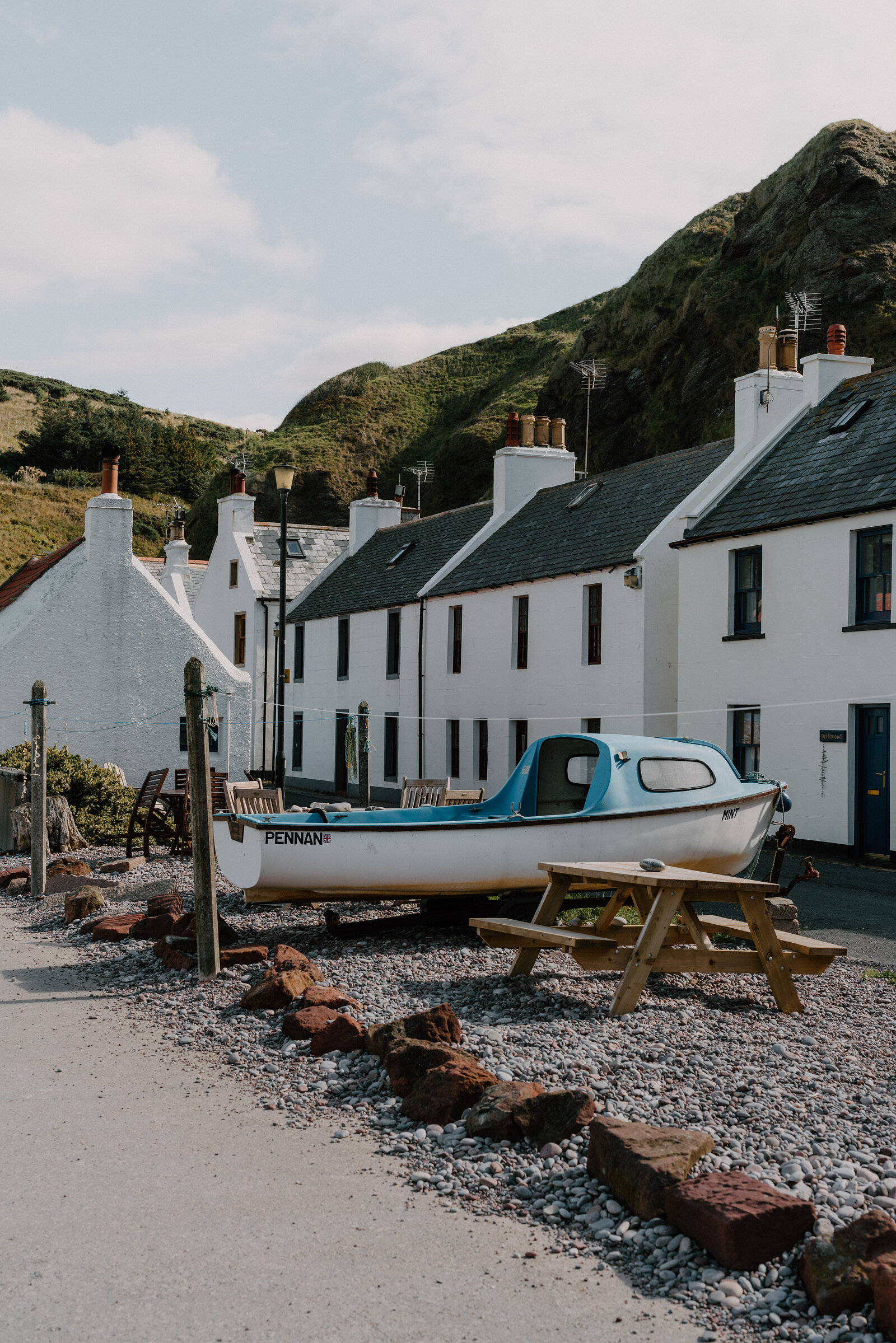 A boat named 'Pennan' sits on the shingle beach in front of the village's white-harled cottages beneath green cliffs.