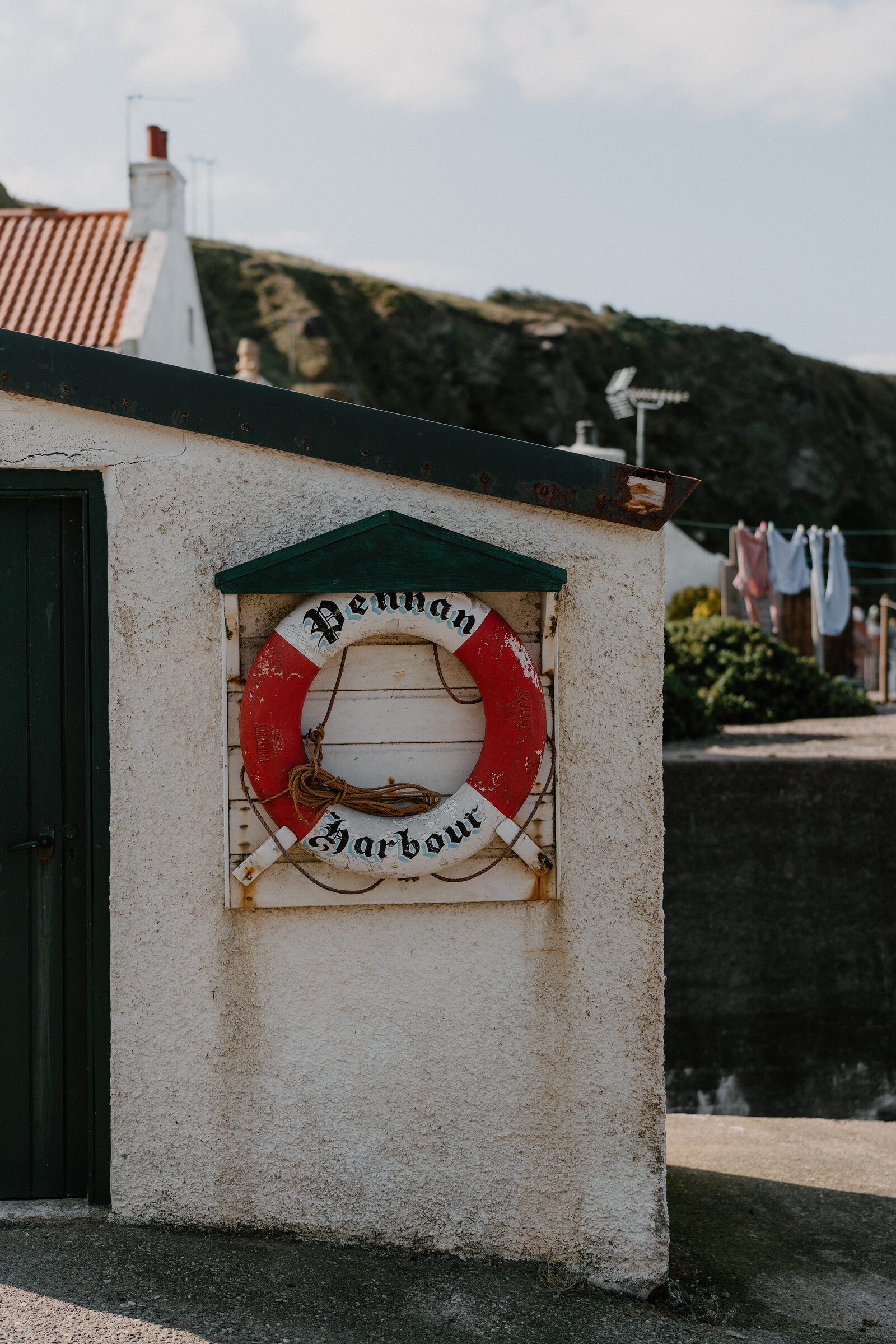 A weathered lifebuoy sign reading 'Pennan Harbour' hangs on a whitewashed building, washing strung out on a line behind.