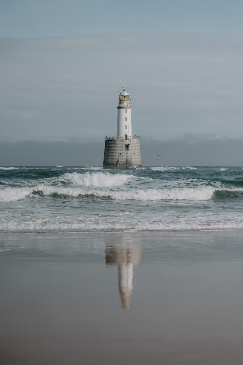 A tall white lighthouse stands offshore in breaking waves off the Aberdeenshire coast, its reflection visible on the wet sand.