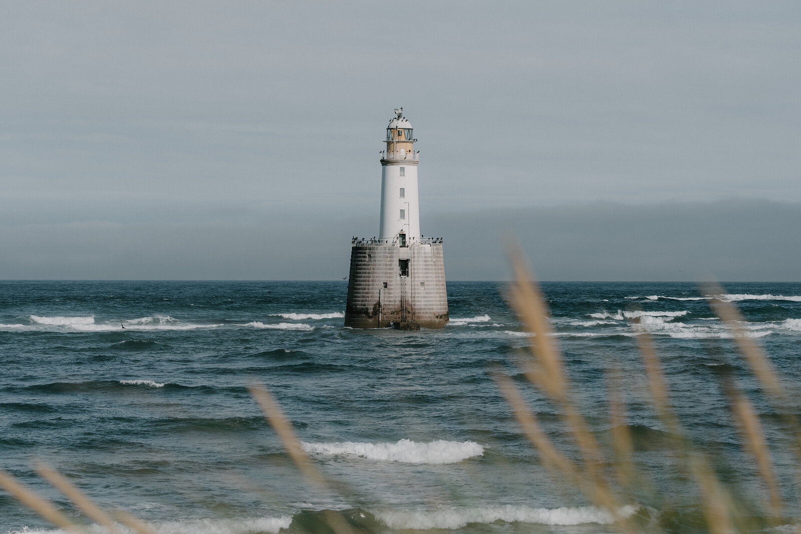 A tall white lighthouse stands offshore in choppy seas off the Aberdeenshire coast, waves breaking around its base.