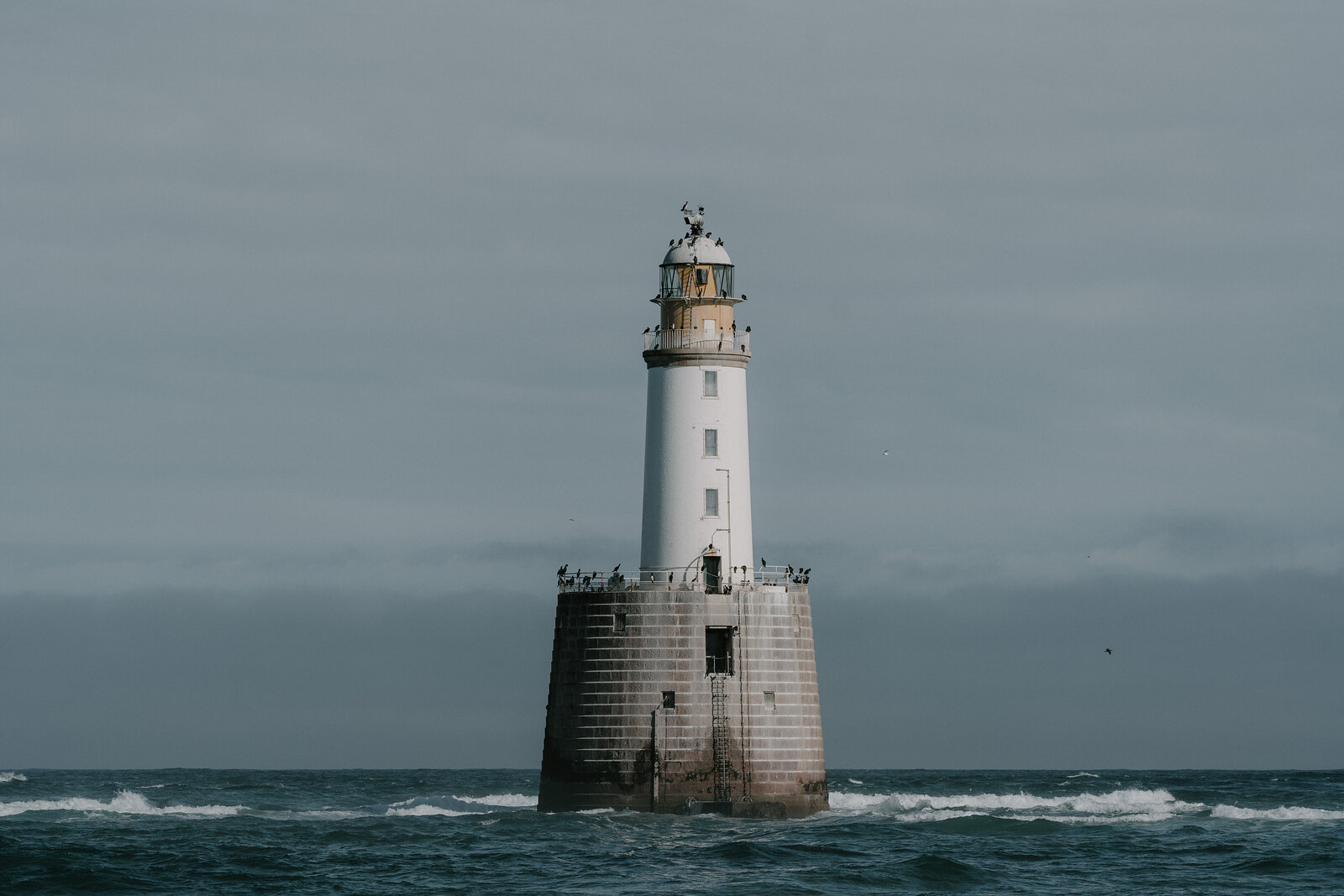 A tall white lighthouse stands offshore in choppy seas off the Aberdeenshire coast, birds gathered around its base.