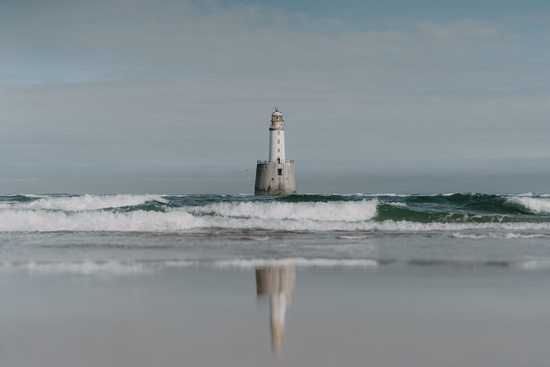 A tall white lighthouse stands offshore amid breaking waves off the Aberdeenshire coast, its reflection visible in the wet sand.
